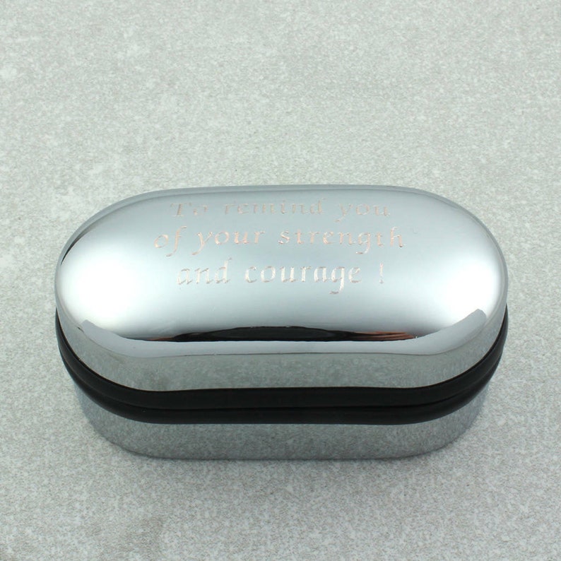 May include: A silver oval-shaped box with a black rim. The box has an engraved message that reads "I'm reminded of your strength and courage!"