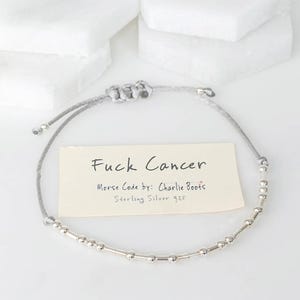 K&ouml;nnte beinhalten: Ein silbernes Morsecode-Armband mit der Aufschrift "Fuck Cancer". Das Armband besteht aus Sterlingsilber 925 und hat ein graues Band. Der Text "Morse Code by: Charlie Boots" ist ebenfalls sichtbar.