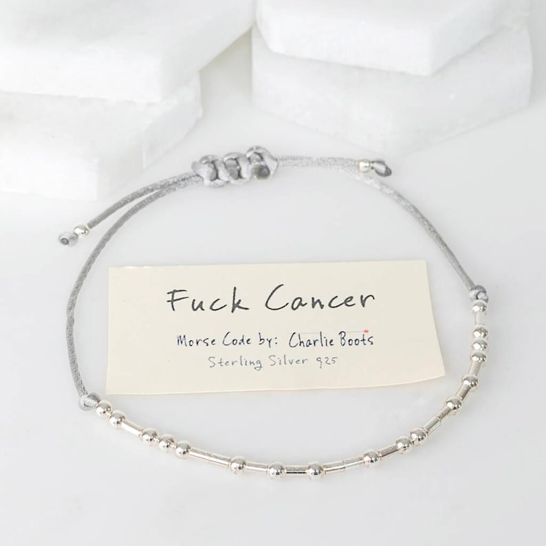 K&ouml;nnte beinhalten: Ein silbernes Morsecode-Armband mit der Aufschrift "Fuck Cancer". Das Armband besteht aus Sterlingsilber 925 und hat ein graues Band. Der Text "Morse Code by: Charlie Boots" ist ebenfalls sichtbar.