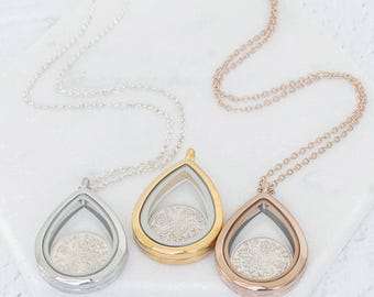 May include: Three teardrop-shaped pendant necklaces in silver, gold, and rose gold. Each pendant features a clear glass front and back, showcasing a coin. The necklaces rest on a white, hexagonal surface.