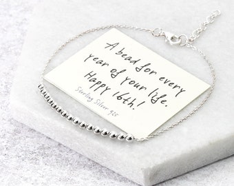 May include: A sterling silver bracelet with a row of small, round beads. A card with the text "A bead for every year of your life. Happy 16th! Sterling Silver 925" is placed behind the bracelet on a marble surface.
