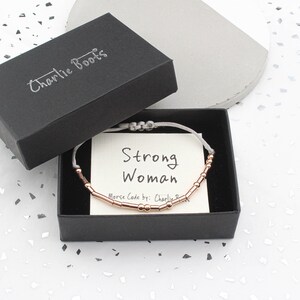 May include: A gray box with a black lid that says "Charlie Boots" on it. The box contains a gray string bracelet with rose gold beads that spell out "Strong Woman" in Morse code. The card inside the box says "Morse Code by: Charlie"