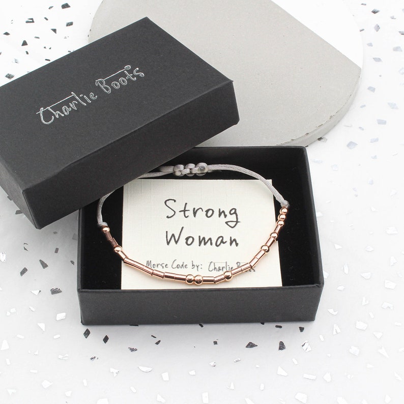 May include: A gray box with a black lid that says "Charlie Boots" on it. The box contains a gray string bracelet with rose gold beads that spell out "Strong Woman" in Morse code. The card inside the box says "Morse Code by: Charlie"