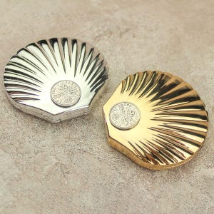 May include: Two shell-shaped trinket boxes, one silver and one gold, each with a coin in the centre. The boxes have a ridged design, resembling a scallop shell. The gold box has a shiny finish, while the silver box has a matte finish.