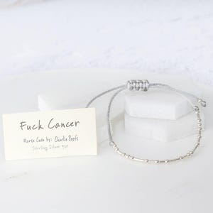 K&ouml;nnte beinhalten: Ein silbernes Morsecode-Armband mit grauem Kordel, pr&auml;sentiert mit einer Karte mit der Aufschrift "Fuck Cancer". Die Karte enth&auml;lt auch "Morse Code by: Charlie Boots" und "Sterling Silver 925". Das Armband liegt auf einer wei&szlig;en Oberfl&auml;che.