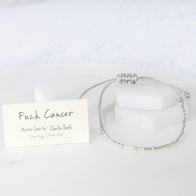 K&ouml;nnte beinhalten: Ein silbernes Morsecode-Armband mit grauem Kordel, pr&auml;sentiert mit einer Karte mit der Aufschrift "Fuck Cancer". Die Karte enth&auml;lt auch "Morse Code by: Charlie Boots" und "Sterling Silver 925". Das Armband liegt auf einer wei&szlig;en Oberfl&auml;che.