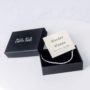 Puede incluir: Una caja negra con las palabras "Charlie Boots" impresas en la parte superior. La caja est&aacute; abierta y contiene una pulsera de plata con una tarjeta que dice "Wonder Woman Morse Code by: Charlie Boots Sterling Silver 925".
