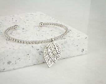 May include: A silver-colored bracelet with a beaded band and a leaf charm. The charm features three stylized leaves with intricate cut-out details. The bracelet is displayed on a textured white surface.