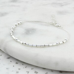 May include: A delicate silver bracelet featuring a series of small, round beads and cylindrical bars strung together. The bracelet has an adjustable chain with a lobster clasp closure. The jewelry is displayed on a white marble surface.