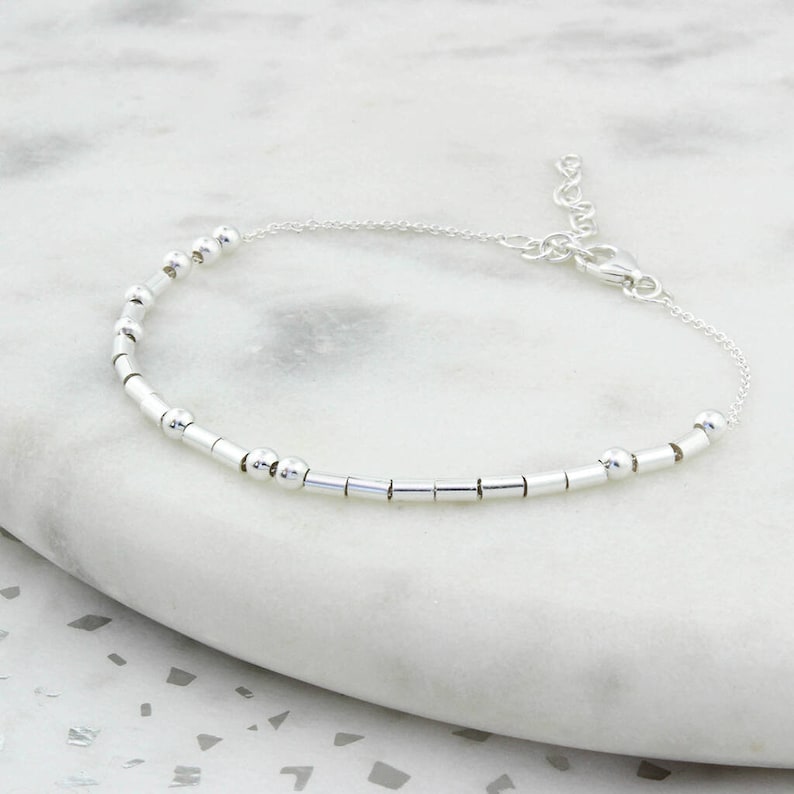 May include: A delicate silver bracelet featuring a series of small, round beads and cylindrical bars strung together. The bracelet has an adjustable chain with a lobster clasp closure. The jewelry is displayed on a white marble surface.