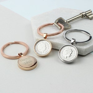 May include: Three silver keyrings with a coin charm. The coin charms feature a profile of Queen Elizabeth II and the text "ELIZABETH II D.G. REG. F.D." The keyring on the left has a rose gold finish and the coin charm has the text "Happy 18th".