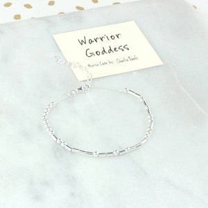 Peut inclure: Un bracelet argenté composé de petites perles rondes et de perles rectangulaires plus longues. Le bracelet est doté d'une fermeture à fermoir. Le bracelet est présenté sur une surface blanche avec une carte qui indique "Warrior Goddess Morse Code by: Charlie Boots".
