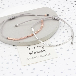 Puede incluir: Dos pulseras con cordón gris y cuentas plateadas y de oro rosa. Las pulseras deletrean "Strong Woman" en código Morse. La tarjeta dice "Strong Woman Morse Code by: Charlie Boots."