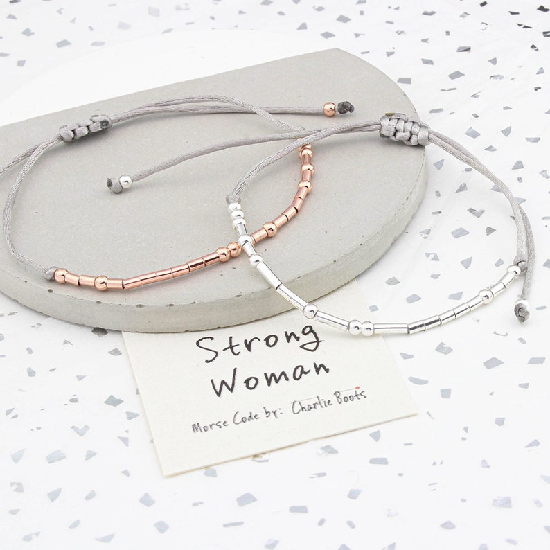 May include: Two bracelets with gray string and silver and rose gold beads. The bracelets spell out "Strong Woman" in Morse code. The card reads "Strong Woman Morse Code by: Charlie Boots."