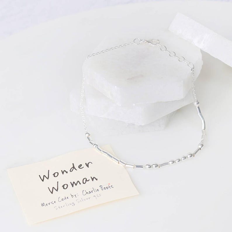 Puede incluir: Pulsera de plata con una cadena delicada y peque&ntilde;as cuentas de plata. La pulsera deletrea "Wonder Woman" en c&oacute;digo Morse. El texto en la tarjeta dice "Wonder Woman Morse Code by: Charlie Boots Sterling Silver $25".