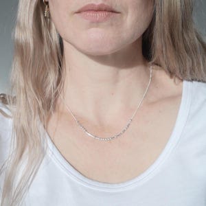 以下が含まれることがあります： 小さなビーズとバーが等間隔に配置された繊細なシルバーネックレス。白いTシャツの上から着用されています。シンプルでエレガントなジュエリーです。