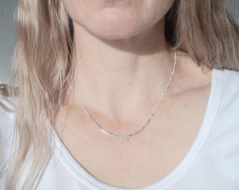 May include: A delicate silver necklace with a series of small, evenly spaced beads and bars. The necklace is worn against a white t-shirt. The necklace is a simple, elegant piece of jewelry.
