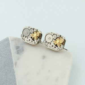 May include: A pair of silver cufflinks with a gold bee design. The cufflinks are made from repurposed watch parts and feature the text "D. SU" and "11CC".
