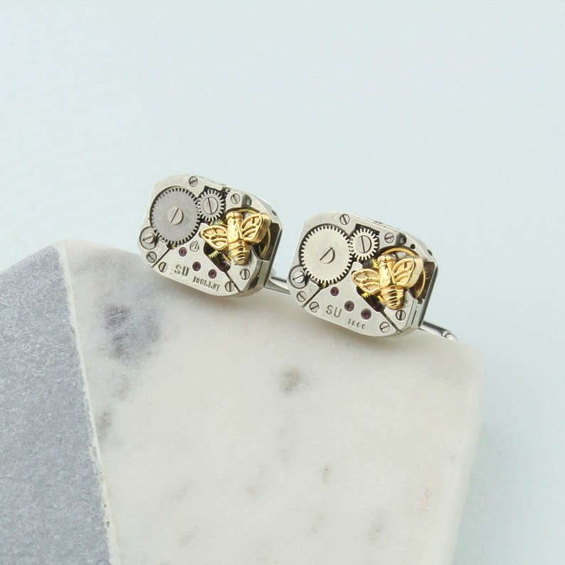 May include: A pair of silver cufflinks with a gold bee design. The cufflinks are made from repurposed watch parts and feature the text "D. SU" and "11CC".