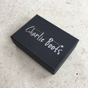 K&ouml;nnte beinhalten: Eine schwarze rechteckige Schachtel mit dem Schriftzug "Charlie Boots" in silberner Schrift. Die Schachtel ist geschlossen und liegt auf einer hellen, strukturierten Oberfl&auml;che. Die Schachtel scheint aus Pappe zu sein.