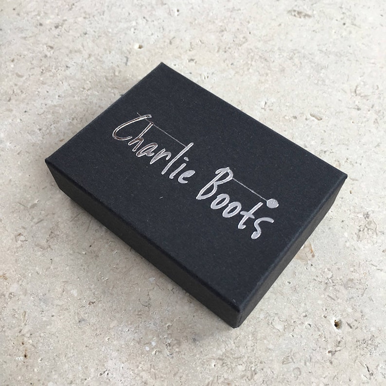 K&ouml;nnte beinhalten: Eine schwarze rechteckige Schachtel mit dem Schriftzug "Charlie Boots" in silberner Schrift. Die Schachtel ist geschlossen und liegt auf einer hellen, strukturierten Oberfl&auml;che. Die Schachtel scheint aus Pappe zu sein.