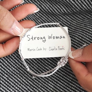 May include: A gray string bracelet with silver beads that spells out "Strong Woman" in Morse code. The bracelet is tied with a knot and has a tag that says "Morse Code by: Charlie Boots".