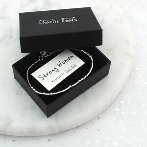 May include: A silver bracelet with a Morse code design, presented in a black box. The box is open, revealing a card that reads "Strong Woman." The brand name "Charlie Boots" is printed on the box. The bracelet is on a white marble surface.