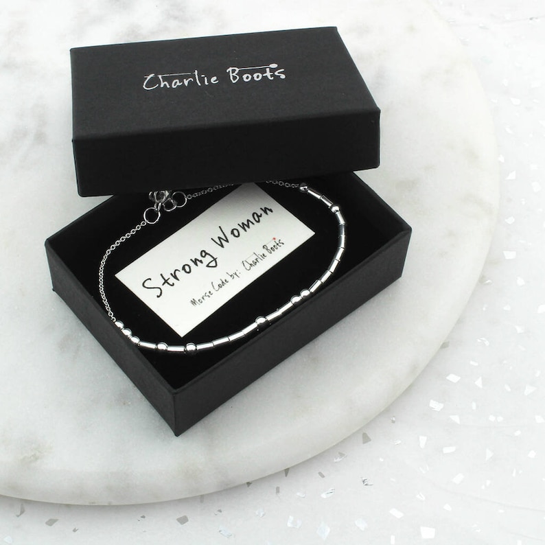 May include: A silver bracelet with a Morse code design, presented in a black box. The box is open, revealing a card that reads "Strong Woman." The brand name "Charlie Boots" is printed on the box. The bracelet is on a white marble surface.