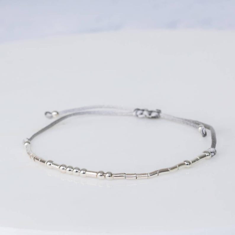 Puede incluir: Un brazalete de cuentas plateadas con un cord&oacute;n gris. El brazalete tiene una mezcla de cuentas redondas y cil&iacute;ndricas.