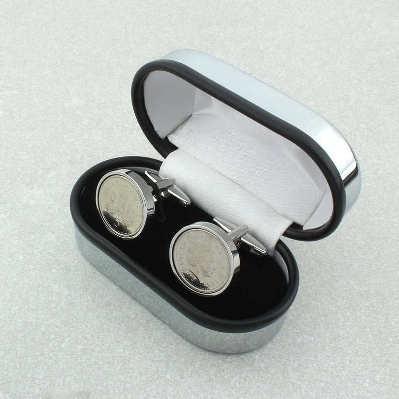 May include: A pair of silver cufflinks in a silver and black box. The cufflinks are round and feature a silver coin design.