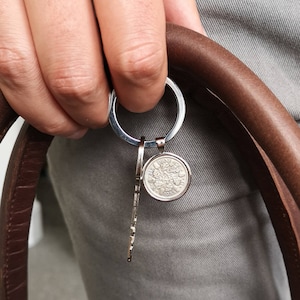 May include: A silver key chain with a round coin charm featuring a design and the year 1953. The keychain is attached to a brown leather strap.