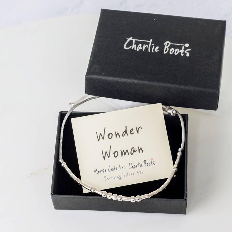 Puede incluir: Un brazalete de plata con un mensaje en c&oacute;digo Morse que dice "Wonder Woman". El brazalete est&aacute; en una caja negra con una tarjeta blanca que dice "Wonder Woman" y "Morse Code by: Charlie Boots Sterling Silver 925".