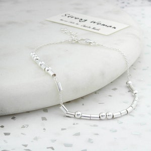 May include: A silver bracelet with a delicate chain and a series of small, cylindrical and spherical beads. The bracelet is on a white surface, and a card with the words "Strong Woman" is in the background.