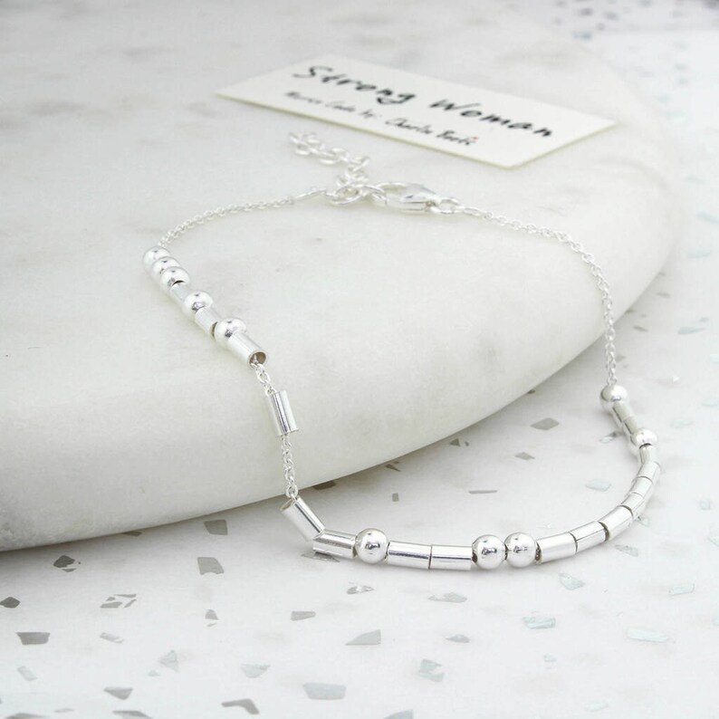 May include: A silver bracelet with a delicate chain and a series of small, cylindrical and spherical beads. The bracelet is on a white surface, and a card with the words "Strong Woman" is in the background.