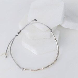 Puede incluir: Un brazalete de cuerda gris con cuentas plateadas y cuentas tubulares plateadas.