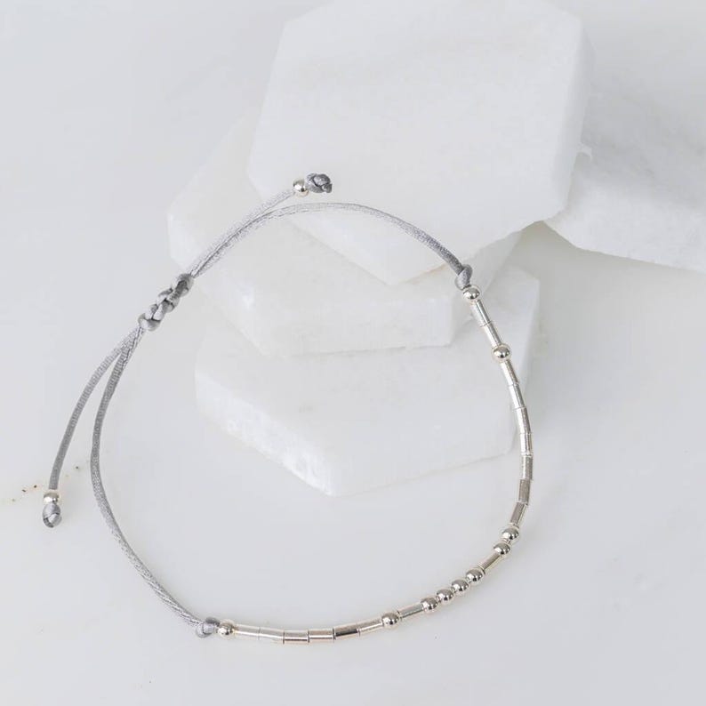 Puede incluir: Un brazalete de cuerda gris con cuentas plateadas y cuentas tubulares plateadas.