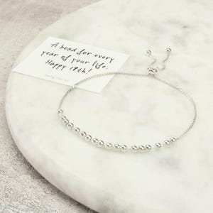 May include: A silver bracelet with small, round beads. A card next to the bracelet reads, "A bead for every year of your life. Happy 18th!" The bracelet is on a white marble surface.