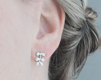May include: Silver stud earrings in a botanical leaf design. The earrings are small and delicate, featuring a cluster of three leaf-shaped elements. The earrings are shown on an ear, highlighting their size and style. The earrings are a simple, elegant accessory.