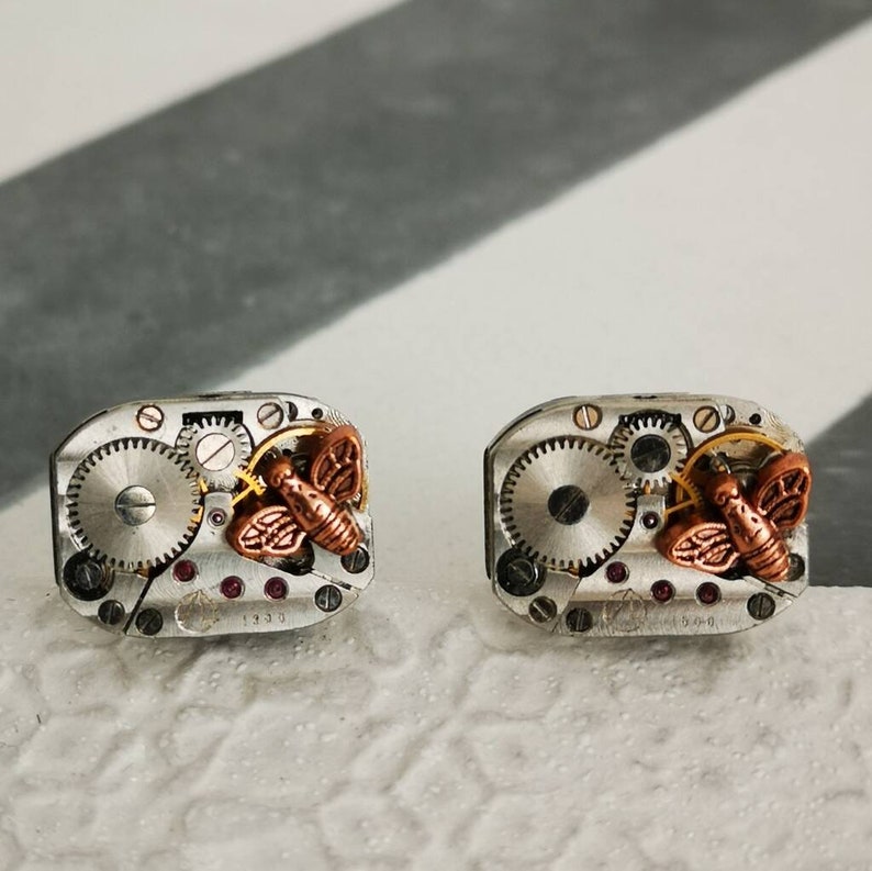 May include: A pair of silver cufflinks with a copper butterfly design and gears. The cufflinks have a vintage look and are a unique accessory.