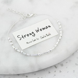 May include: Silver bracelet with small round and rectangular beads. The bracelet has a clasp closure. A card with the text "Strong Woman" and "Morse Code by: Charlie Boots" is in the background.
