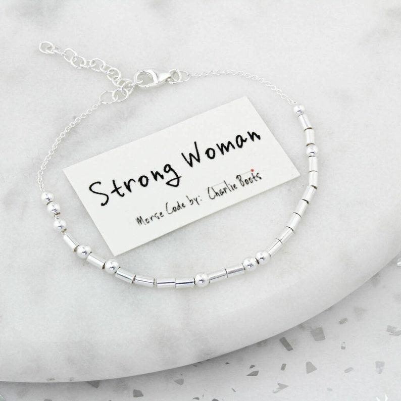 May include: A silver Morse code bracelet with the words "Strong Woman" on a white card. The bracelet features small, cylindrical and spherical beads. The card also includes the text "Morse Code by: Charlie Boots".