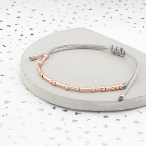 May include: A delicate bracelet with a gray cord and rose gold beads. The bracelet is adjustable and can be worn on any wrist size.