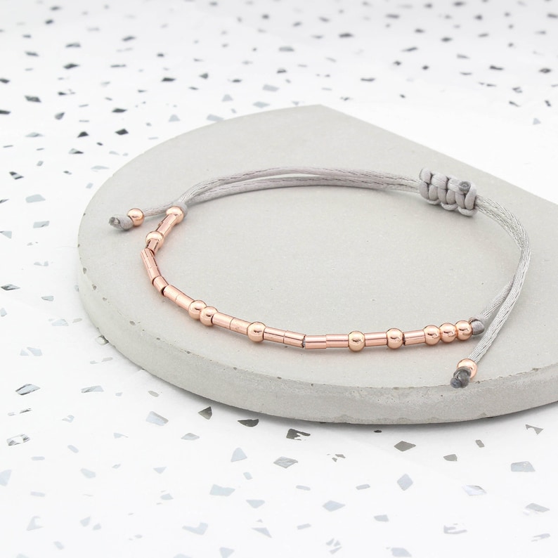 May include: A delicate bracelet with a gray cord and rose gold beads. The bracelet is adjustable and can be worn on any wrist size.
