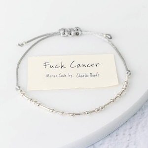 K&ouml;nnte beinhalten: Ein silbernes Perlenarmband mit einem grauen Band. Das Armband zeigt den Text "Fuck Cancer" auf einer kleinen Karte, darunter "Morse Code by: Charlie Boots". Das Armband liegt auf einer wei&szlig;en Oberfl&auml;che.