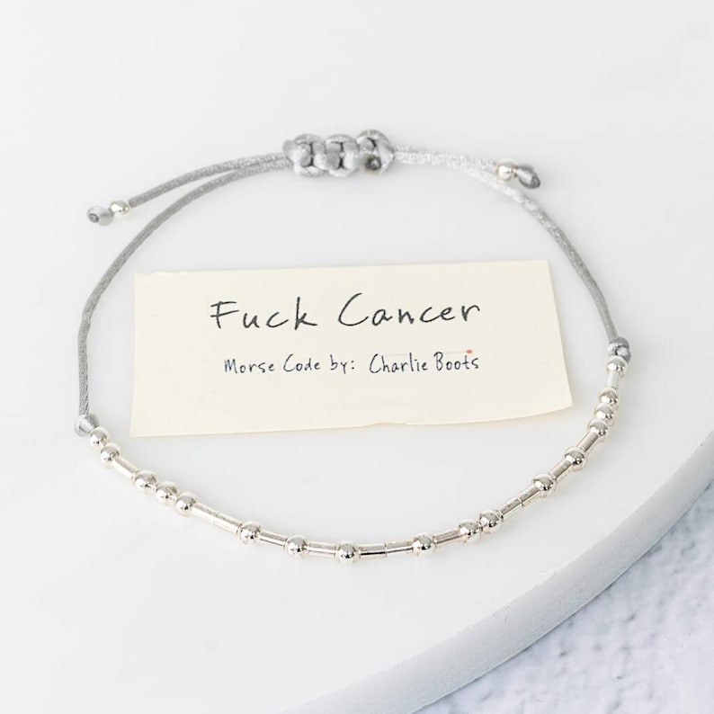 K&ouml;nnte beinhalten: Ein silbernes Perlenarmband mit einem grauen Band. Das Armband zeigt den Text "Fuck Cancer" auf einer kleinen Karte, darunter "Morse Code by: Charlie Boots". Das Armband liegt auf einer wei&szlig;en Oberfl&auml;che.