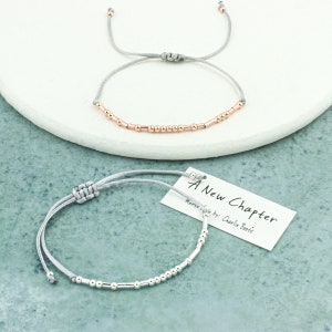 Può includere: Due braccialetti regolabili con cordoncino grigio e perline d'argento e oro rosa. Il braccialetto in basso ha un'etichetta che recita "A New Chapter Morse Code by: Charlie Boots".
