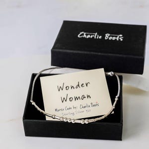 Puede incluir: Una caja negra con un brazalete de plata en su interior. El brazalete est&aacute; hecho de cuentas de plata de ley y est&aacute; grabado con las palabras "Wonder Woman" en c&oacute;digo Morse. La caja tiene una tarjeta blanca en su interior con las palabras "Wonder Woman" y "Morse Code by: Charlie Boots" escritas en ella.