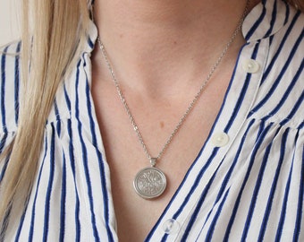May include: A silver-toned pendant necklace with a circular pendant featuring a detailed floral design. The necklace is worn over a white and blue striped blouse. The chain is delicate and the pendant is centered.