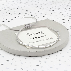 May include: A gray string bracelet with silver beads spelling out "Strong Woman" in Morse code. The bracelet is on a gray card with the text "Strong Woman" and "Morse Code by: Charlie Boots".