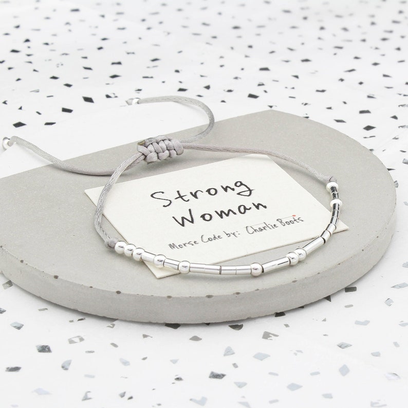 May include: A gray string bracelet with silver beads spelling out "Strong Woman" in Morse code. The bracelet is on a gray card with the text "Strong Woman" and "Morse Code by: Charlie Boots".
